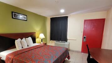 A hotel room with a bed, a desk, a chair, a lamp, a window with curtains, a door, and a picture on the wall.
