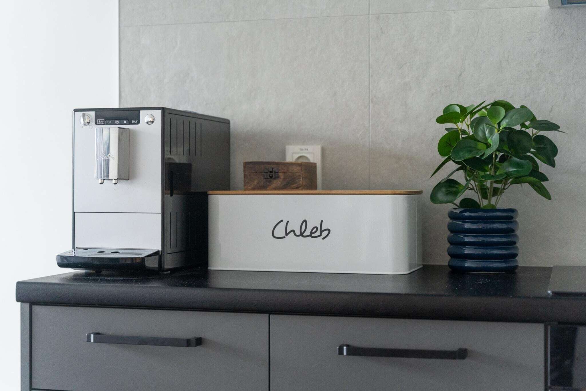 Una cucina con una macchina per il caffè, una pianta in un vaso blu e un mobiletto a cassetti con l'etichetta 'Chleb'.