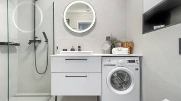 A modern bathroom with a glass-enclosed shower, a built-in sink with a mirror, a washing machine, and a small shelf with toiletries.