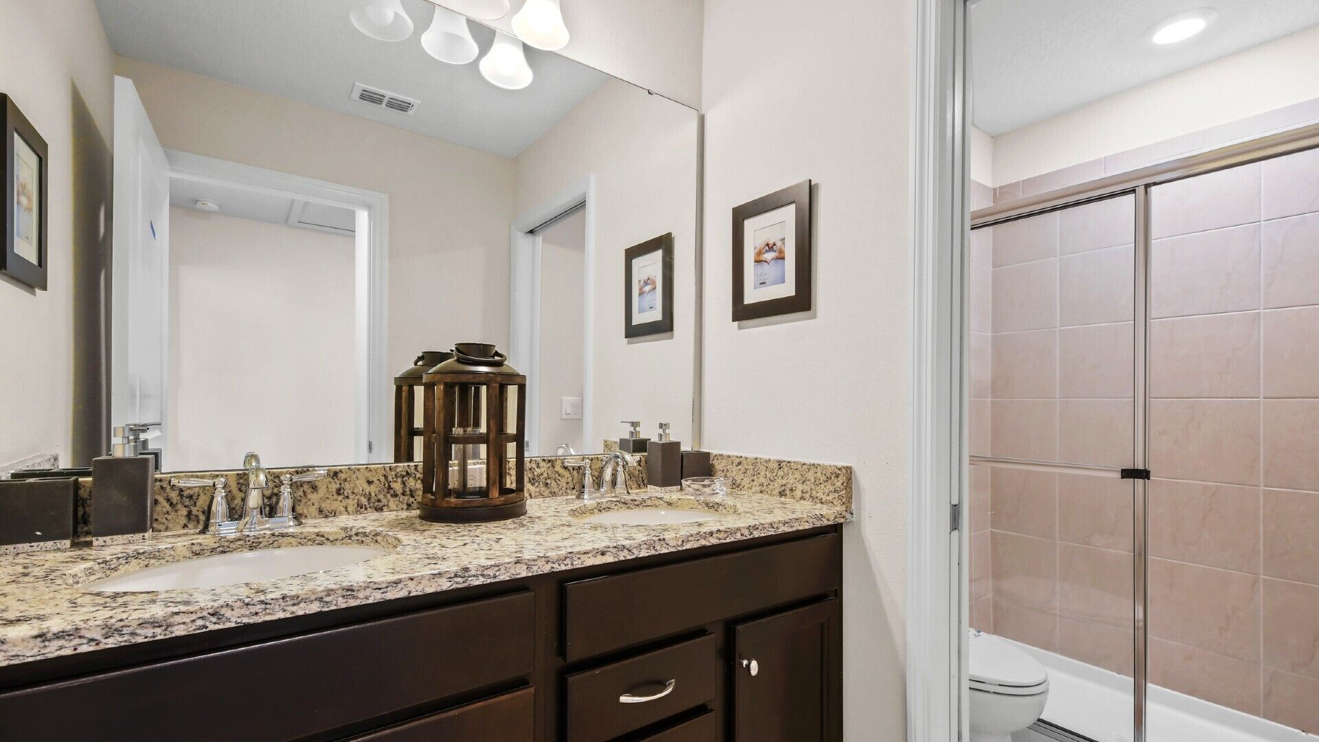 A bathroom with a granite countertop, a large mirror, a lantern, framed pictures, and a shower area.