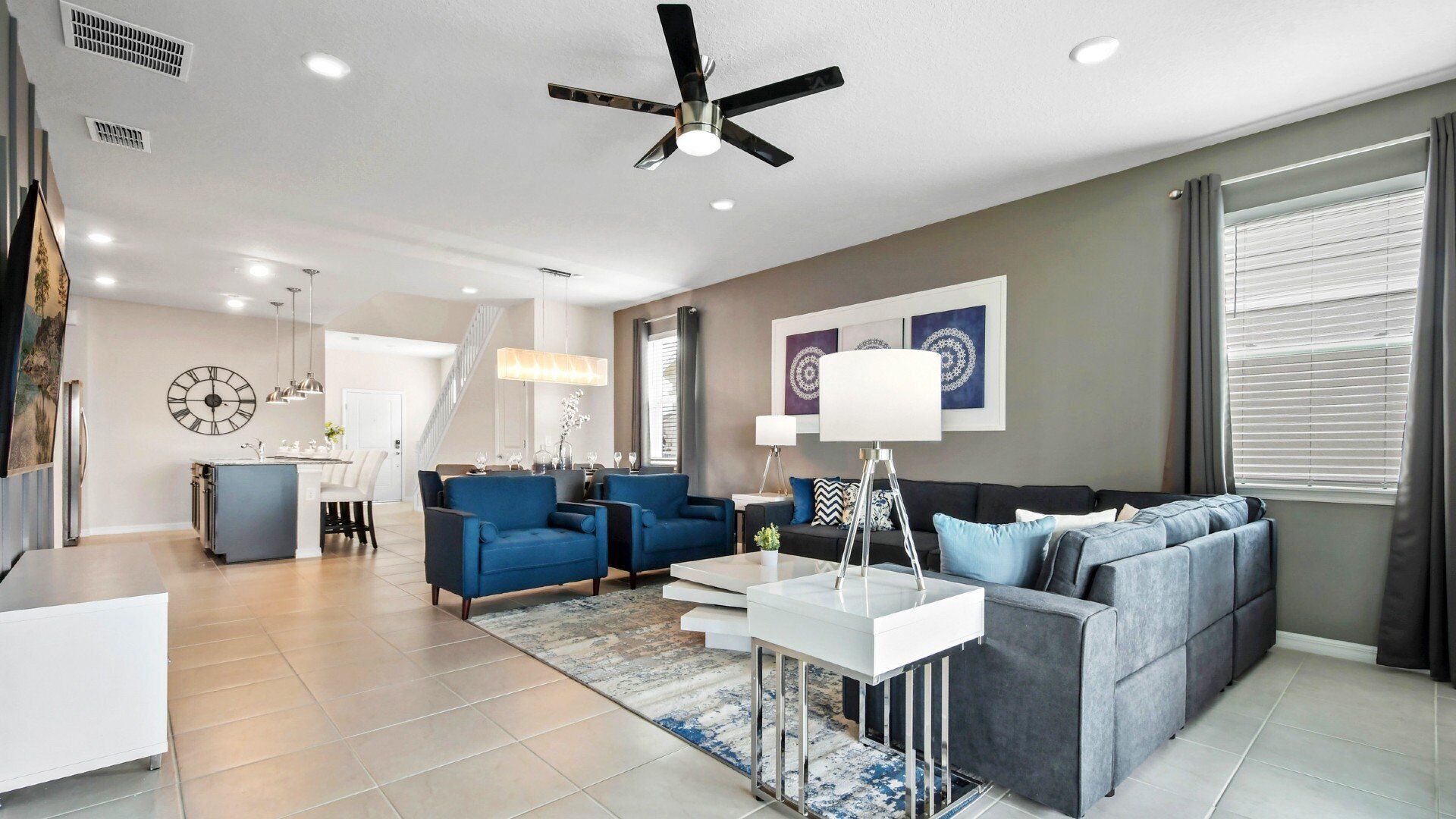 Un salon moderne avec un canapé gris, des fauteuils bleus, une table basse blanche et un ventilateur de plafond.