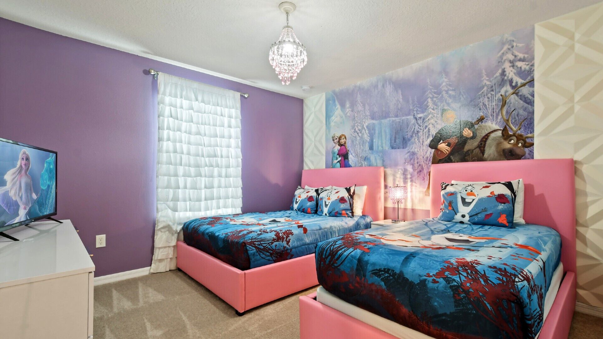 A bedroom with two pink beds, a television on a white desk, and a chandelier.