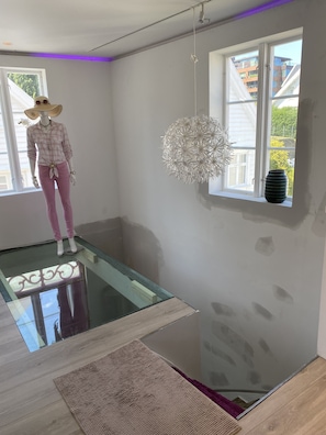 A room with a glass floor, a mannequin, a hanging light fixture, and a window with a view of buildings. - Sweat Home Old Stavanger (Stavanger)