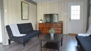 A living room with a dark sofa, a coffee table, a wooden cabinet with a television, and a door. - Cottage with sunset view and much more (Alburgh)