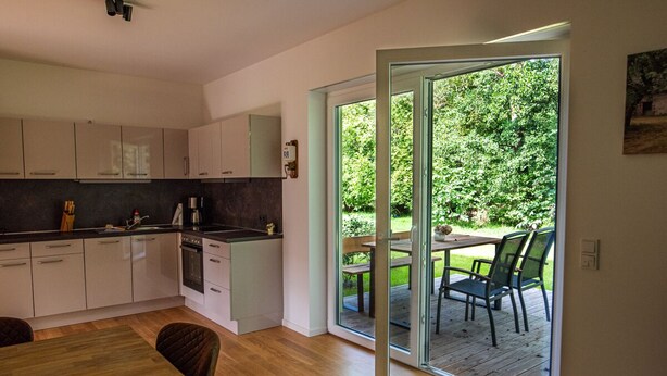 Une cuisine moderne avec des meubles blancs, une table à manger en bois et une porte-fenêtre coulissante donnant sur une terrasse extérieure avec une table et des chaises.