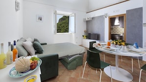 Habitació petita i funcional amb llit, taula amb esmorzar, finestra amb vista a vegetació i petita cuina equipada.