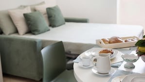 Taula de petit esmorzar ben ordenada amb pastissos, una gerra i una tassa, davant d'una habitació moderna amb un llit i un sofà verd.