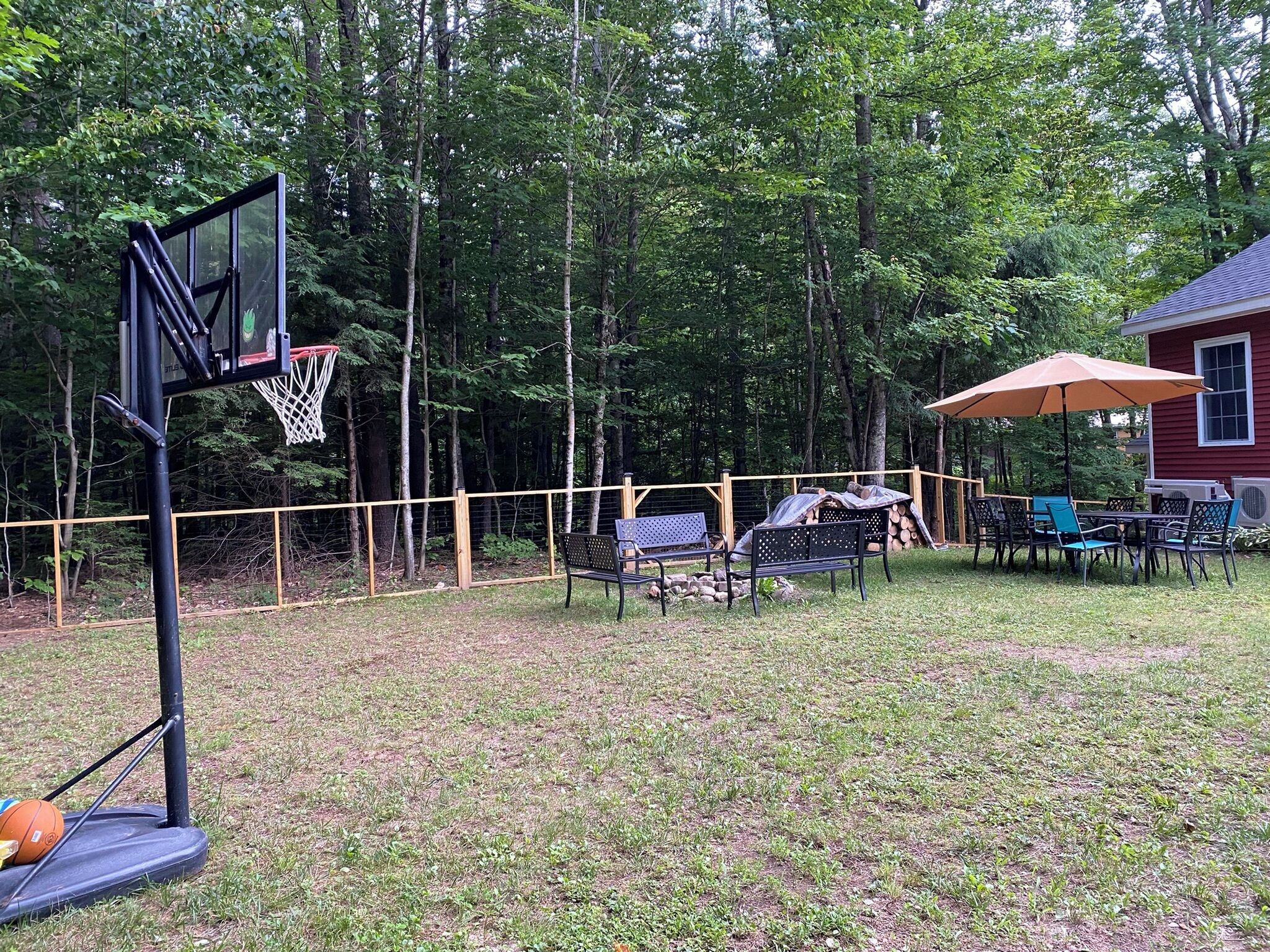 A basketball hoop, a covered patio area with chairs, and a red building in a wooded setting.