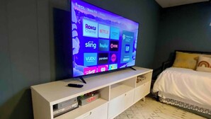 Un dormitorio con una televisión sobre un soporte blanco que muestra diferentes servicios de streaming.