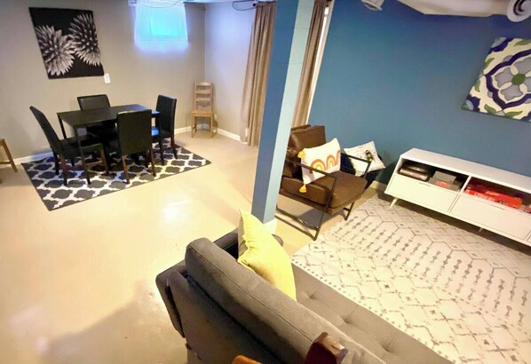 A living room with a grey sofa, a dining table with chairs, a patterned rug, a white cabinet, and a blue wall.