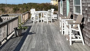 Eine Holzterrasse mit weißen Stühlen und einem Tisch, mit Blick auf einen Strand und einen bewaldeten Bereich.