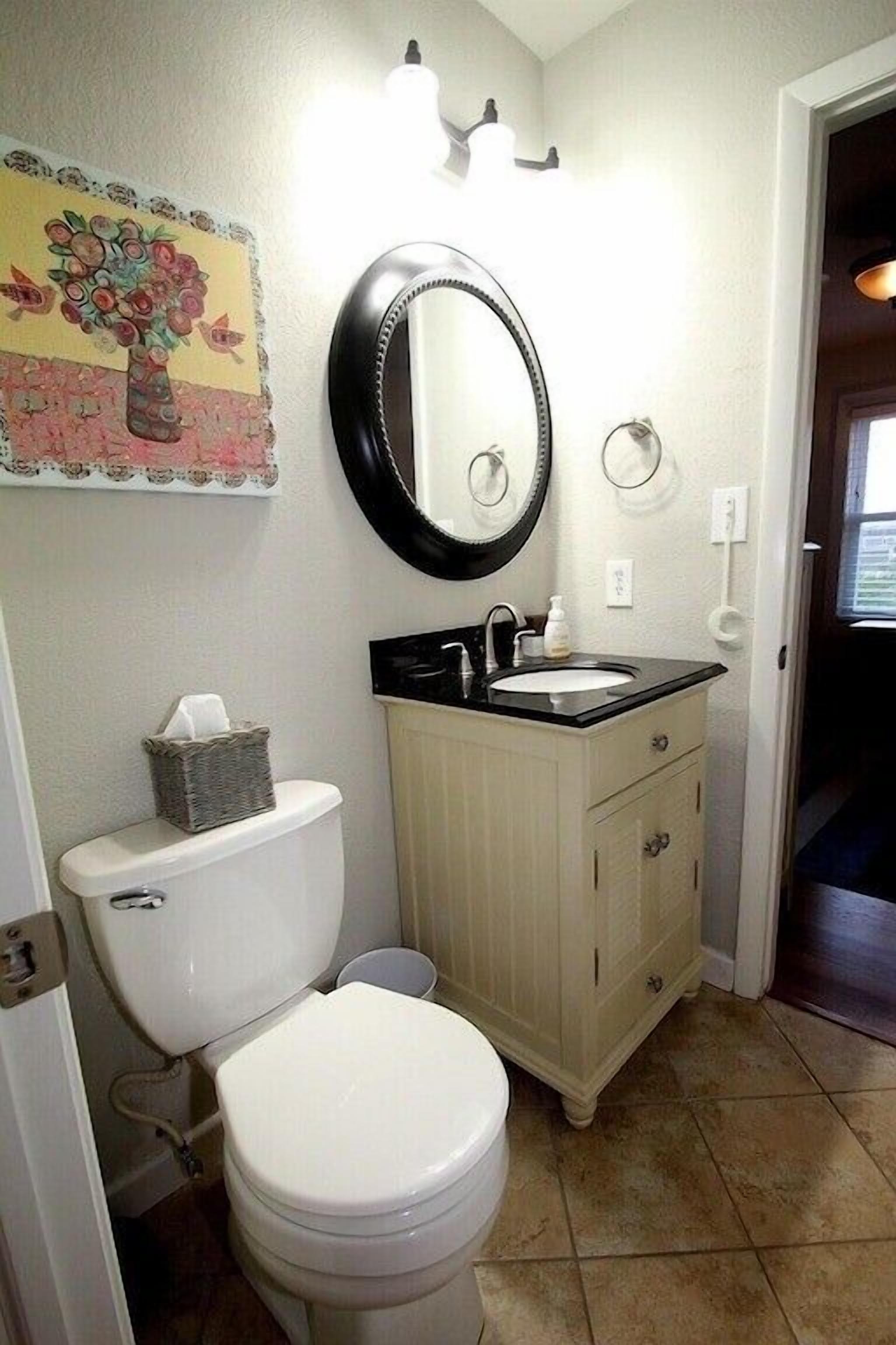 Une salle de bain avec des toilettes, un lavabo et un miroir. Il y a un tableau accroché au mur et un panier de mouchoirs sur les toilettes.