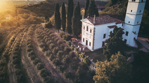 Une vue aérienne d’un bâtiment blanc surmonté d’une tour, entouré d’arbres et situé dans un paysage vallonné.