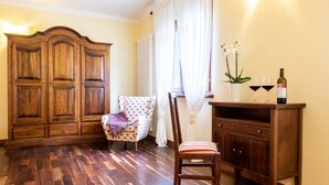 Une pièce avec un plancher en bois, une grande armoire, une chaise recouverte d’un tissu à motifs, une petite table avec deux verres à vin et une bouteille, et un pot d’orchidée.
