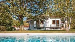 Une maison à deux étages avec un balcon, une piscine au premier plan et des arbres en arrière-plan.
