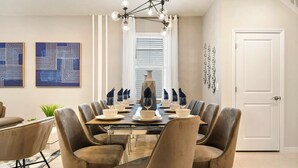 Un coin repas avec une longue table dressée pour six personnes, entourée de chaises rembourrées. Deux tableaux abstraits ornent le mur, un grand vase est posé sur la table, et une porte est dotée de poignées décoratives.