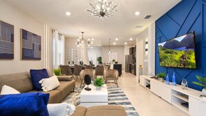 Un salon moderne avec un canapé, une table basse et un téléviseur. On aperçoit un coin repas avec des chaises et, au fond, une cuisine.