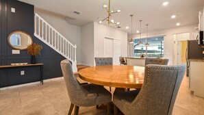 A dining area with a round wooden table and upholstered chairs, a staircase with white railings, and a kitchen in the background.
