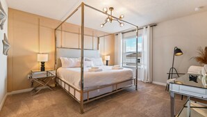 A four-poster bed with white linens, bedside lamps, a desk with a lamp, and a view of the outside through the window.