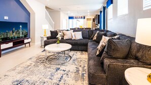 A modern living room with a sectional sofa, a coffee table, and a large flat-screen TV displaying a cityscape. - Last Minute Disney Getaway! Luxury Villa with Private Pool & Game Room. Book Now! #87165 (Orlando)