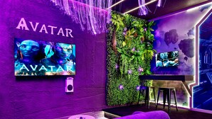 Huoneessa on seinälle asennettu televisio, jossa näkyy elokuva »Avatar», vihreä kasviseinä, violetti sohva ja baaritiski.