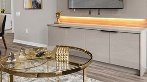 A modern living room with a glass coffee table, a flat-screen TV mounted on a wooden shelf, and a decorative plant. - Last Minute Disney Getaway! Luxury Condo with Private Balcony. Book Now! #89639 (Orlando)