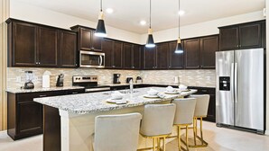 A modern kitchen with dark wood cabinets, a stainless steel refrigerator, a built-in microwave, and a granite countertop. There are bar stools and a coffee maker on the island. - Last Minute Disney! Private Pool, Hot Tub, Game Room & Themed Bedrooms. Book Now! #90909 (Orlando)