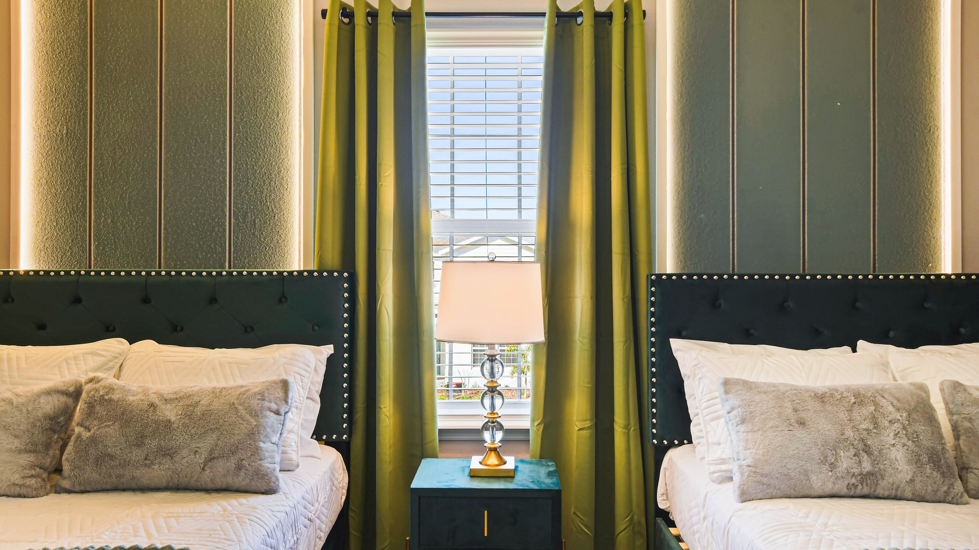 Habitación de hotel con dos camas, cortinas verdes, una mesita con lámpara y una ventana con persianas.