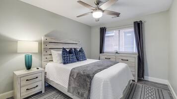 A bedroom with a bed, a nightstand, a lamp, a ceiling fan, a window with curtains, and a view of the outside.