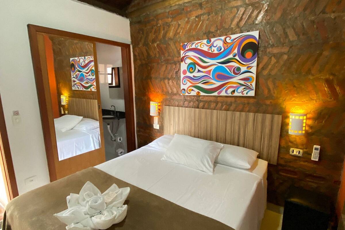 A hotel room with a large bed, a wooden ceiling, a brick wall, and a colorful painting.