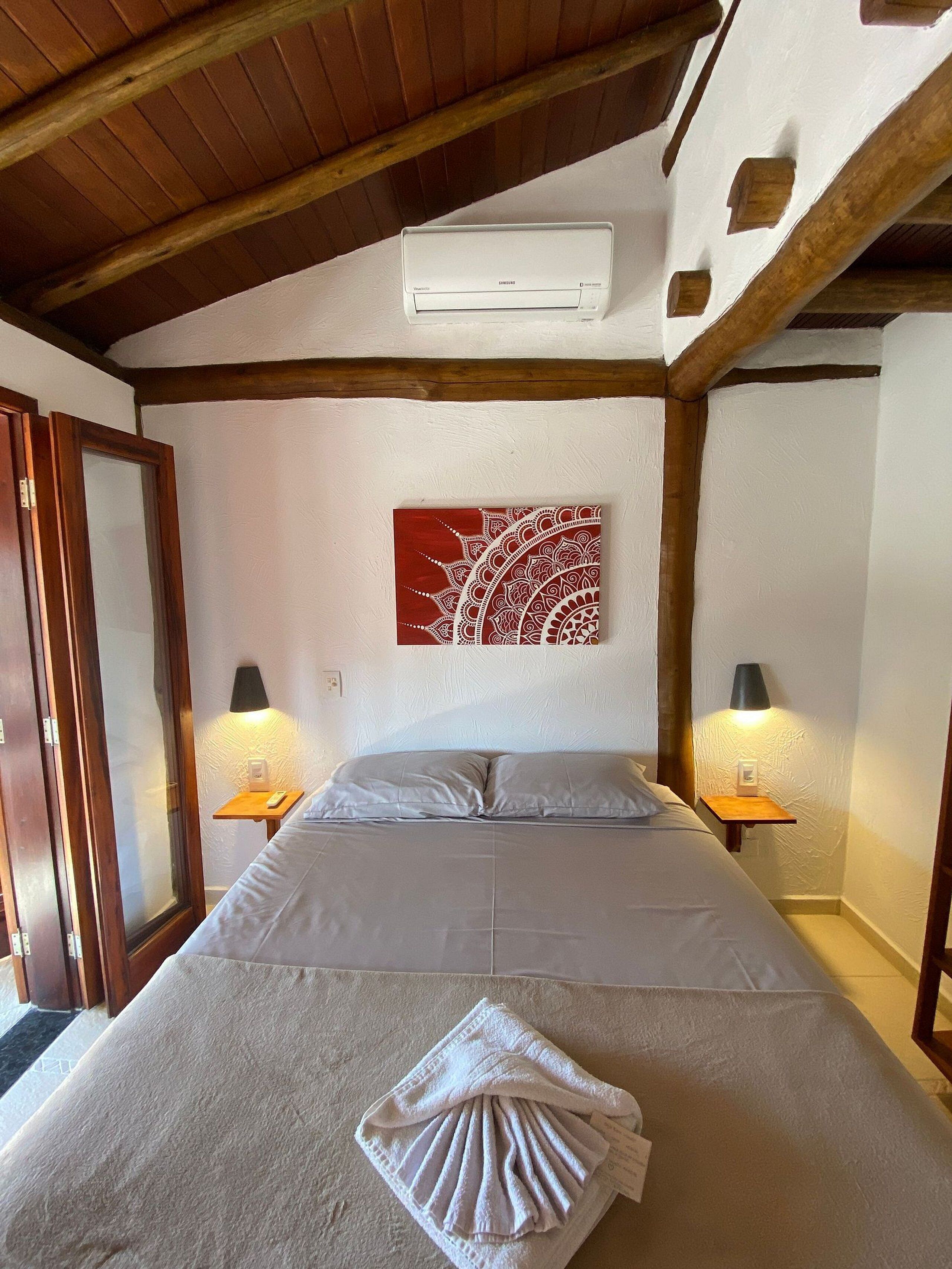 A bedroom with a bed, two bedside lamps, a wall-mounted air conditioner, and a decorative wall piece.