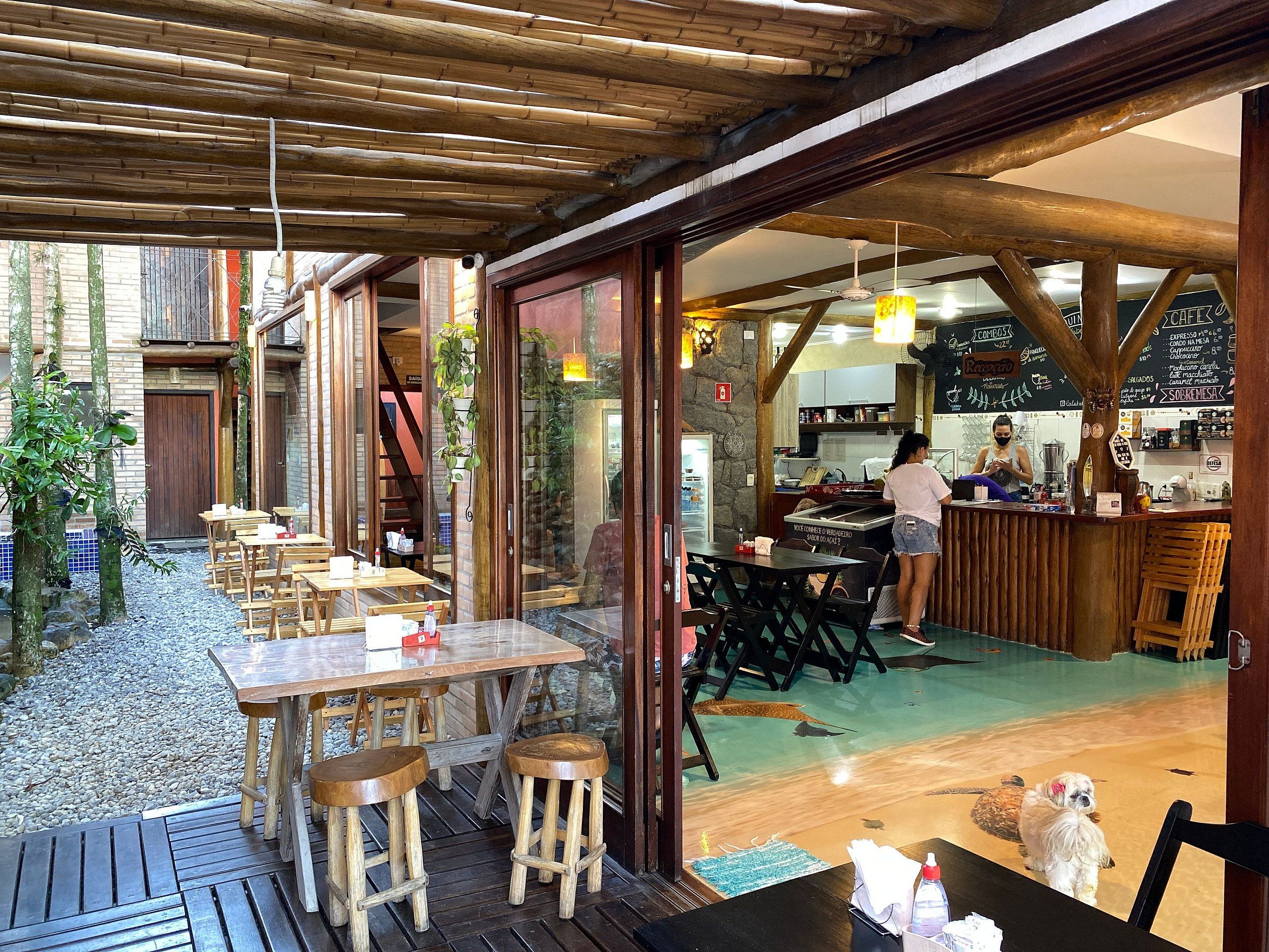 An outdoor dining area with wooden tables and stools, a stone wall, and a view of the kitchen area.