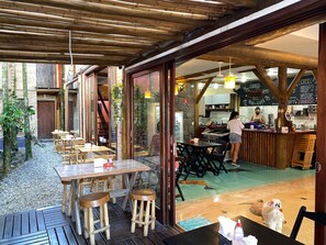 An outdoor dining area with wooden tables and stools, a stone wall, and a view of the kitchen area. - Lalabella Itagua (Ubatuba)