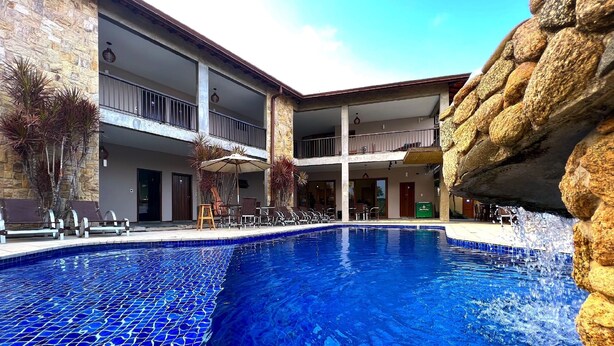 Una piscina con bordes azules de azulejos, rodeada de sillones de descanso y sombrillas, con un muro de piedra y un edificio de varios niveles al fondo.