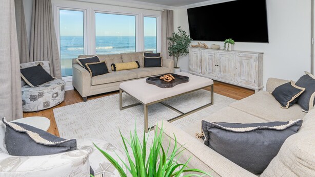 A living room with a sofa, armchairs, a coffee table, and a TV. There are large windows with a view of the ocean.