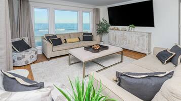 A living room with a sofa, armchairs, a coffee table, and a TV. There are large windows with a view of the ocean.