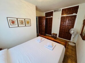 A hotel room with a bed, a wooden wardrobe, a small stool, and a standing fan. - Comfortable | Very quiet area | 2km from Cala Cap Blanc - Punta Estrella (VC)
