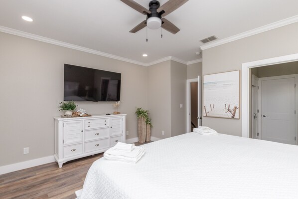 A bedroom with a large bed, a dresser, a ceiling fan, a TV, and a door leading to another room. - Island Escape | Pool Access & Beach Nearby (Corpus Christi)
