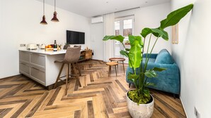 A modern living room with a herringbone wooden floor, a blue sofa, a dining area with a white table and chairs, and a large green plant.