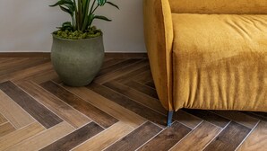 A room with a herringbone wooden floor, a green potted plant, and a mustard-colored sofa against a white wall.