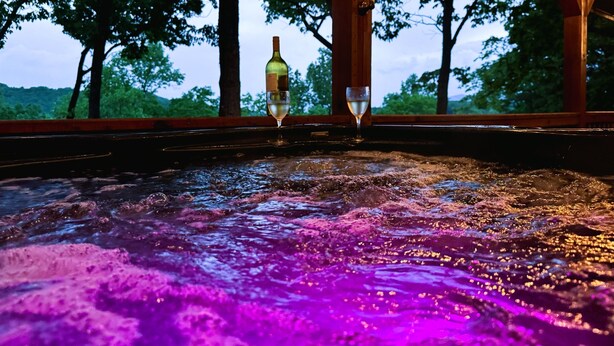 A hot tub with a bottle of wine and a glass, surrounded by trees and a wooden structure. - 🌿 Summer Retreat  *  HotTub  *  Mountain Views  *  2 Masters  *  Pets OK (Ellijay)