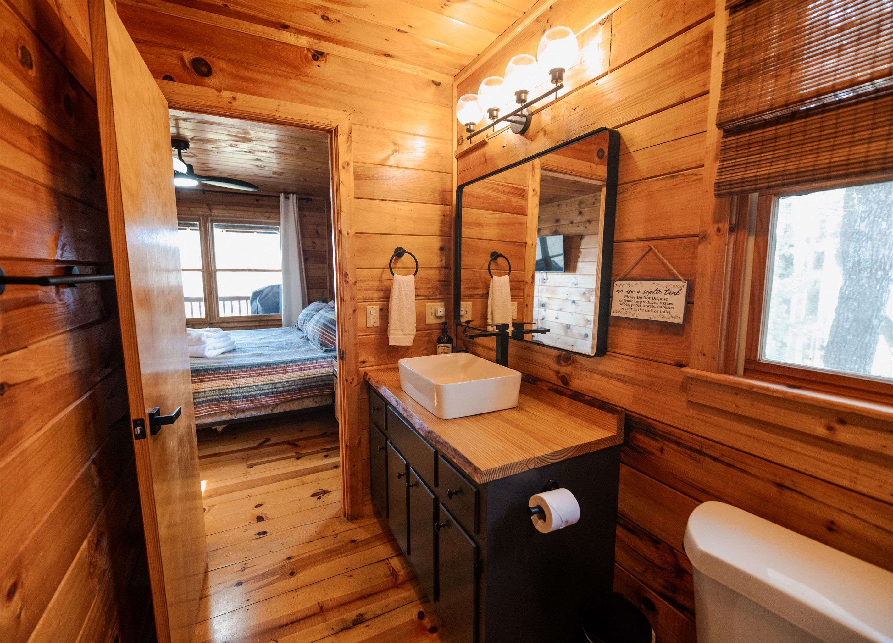 A wooden cabin interior with a bathroom featuring a sink, mirror, and toilet. A bed with a striped blanket is visible in the background.