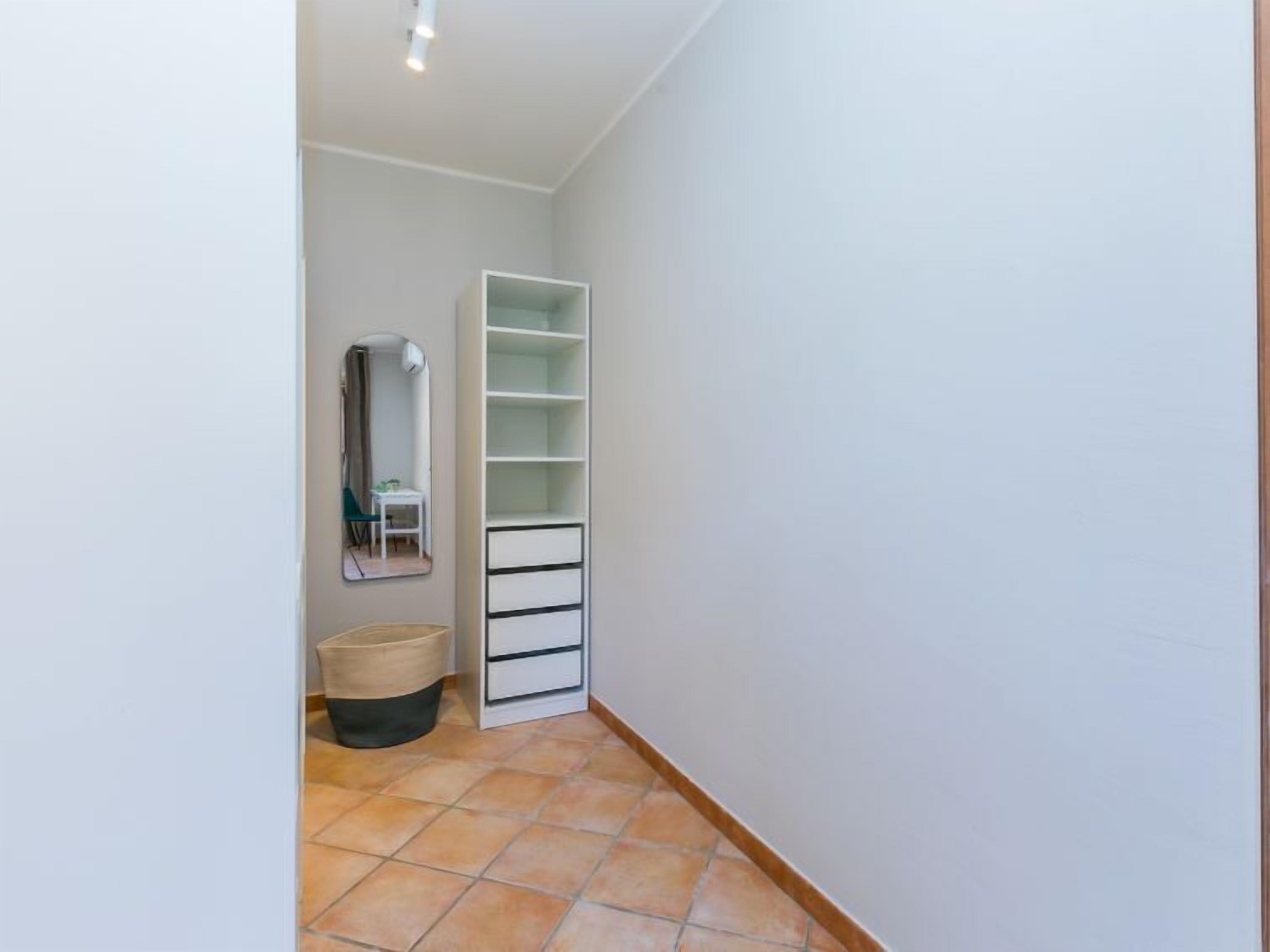 A narrow hallway with a white wall, a tiled floor, a white storage unit with drawers, a round wicker basket, and a mirror on the wall.