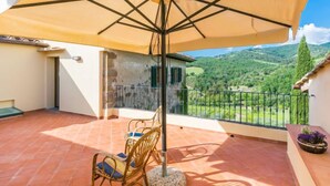 Un balcone con un ombrellone grande, sedie in vimini e vista su colline verdi.
