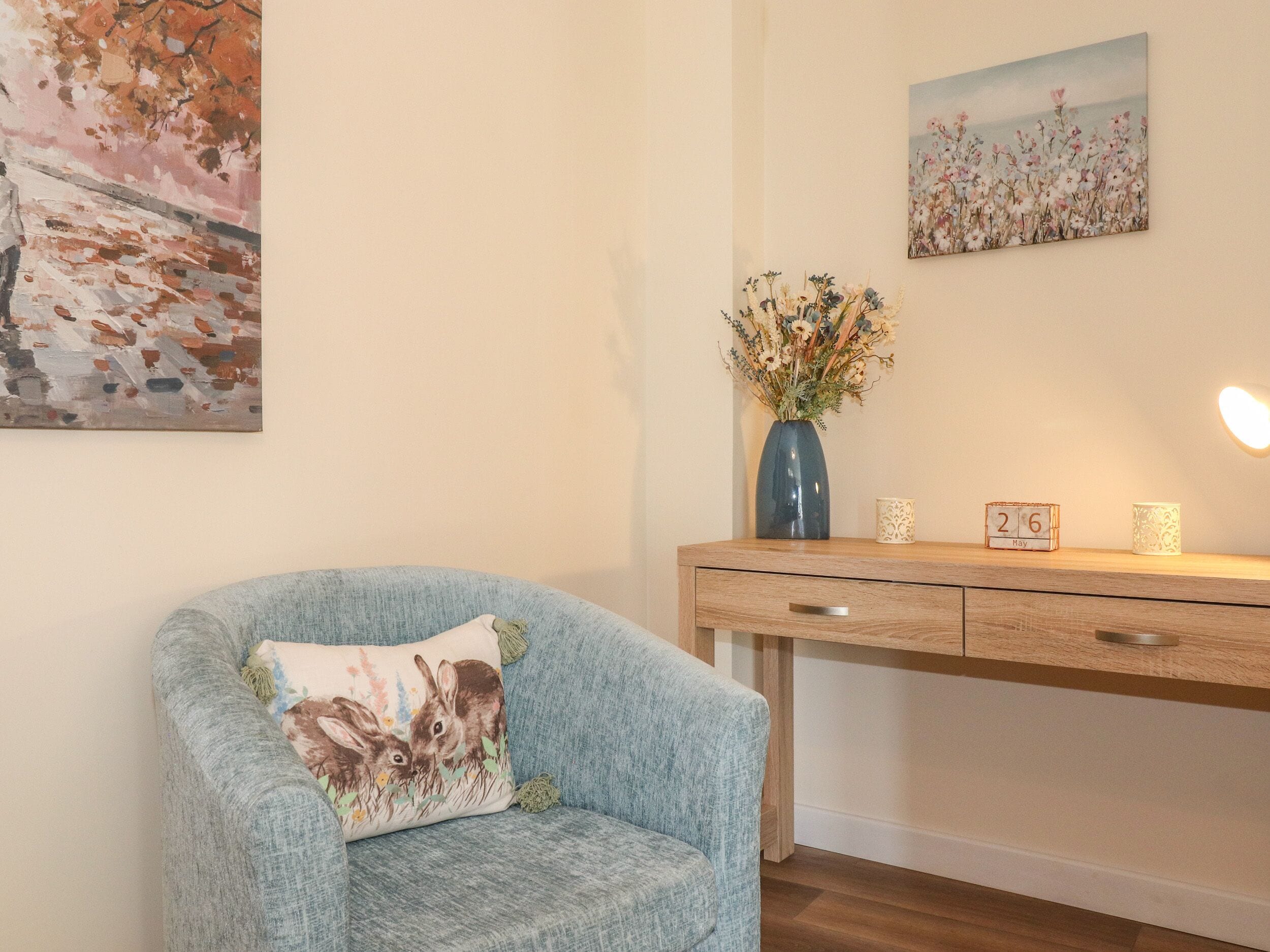 A room with a light blue armchair, a wooden desk, a vase with flowers, and two wall-mounted paintings.