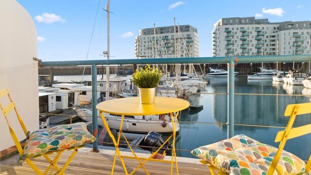 A balcony with yellow chairs and a small round table, overlooking a marina with boats and buildings in the background. - On the Water 4 With Free Parking By My Getaways (Brighton)
