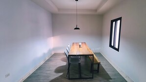 Ein Zimmer mit einem langen Holztisch, zwei Stühlen, einer hängenden Pendelleuchte und einem Fenster mit vertikalen Jalousien.