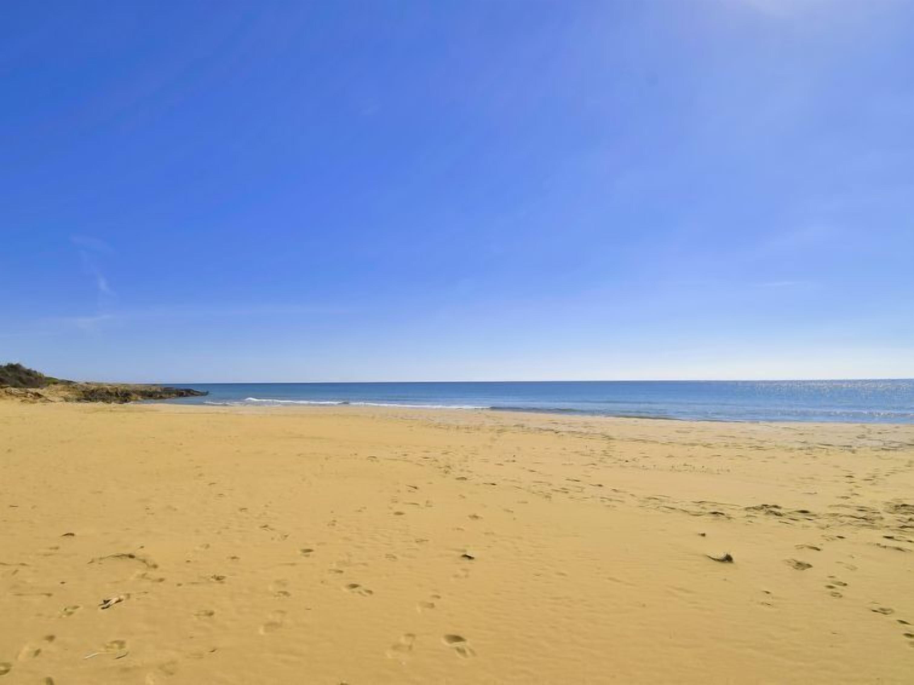 Een breed zandstrand met voetafdrukken die naar het water leiden, onder een heldere blauwe lucht.