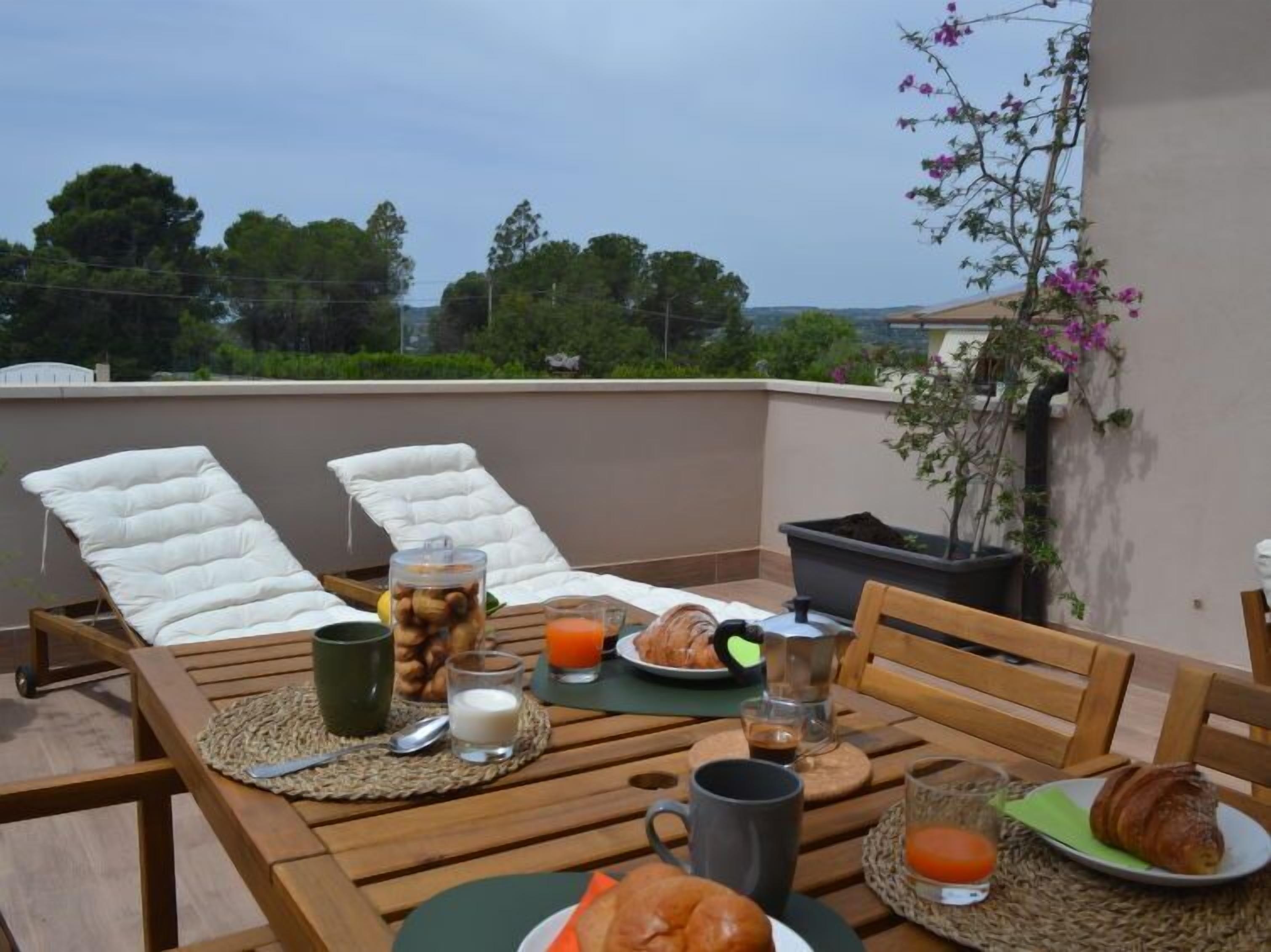 A rooftop terrace with two lounge chairs, a table set with breakfast items including croissants, juice, and coffee, and a view of trees and buildings in the background.
