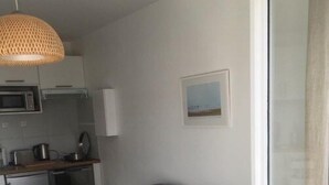 A compact bedroom with a bed, a small bedside table, a chair, a kitchenette with a microwave, and a hanging light fixture. - Un Balcon sur la Mer - Appt à 30m de la plage (Saint-Aubin-sur-Mer)
